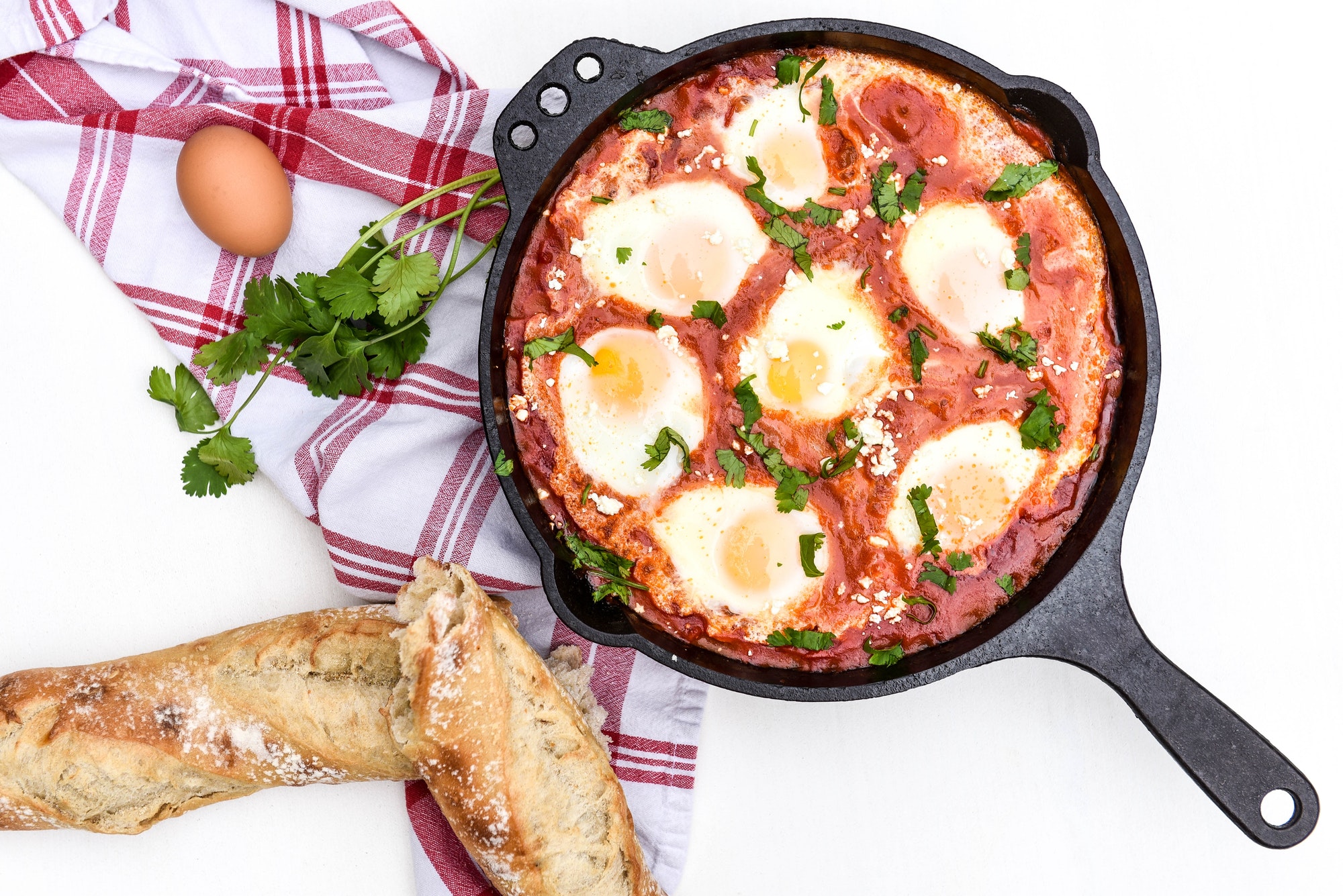 Flat lay of a shakshuka dish in a cast iron skillet next to a torn baguette and ingredients on a